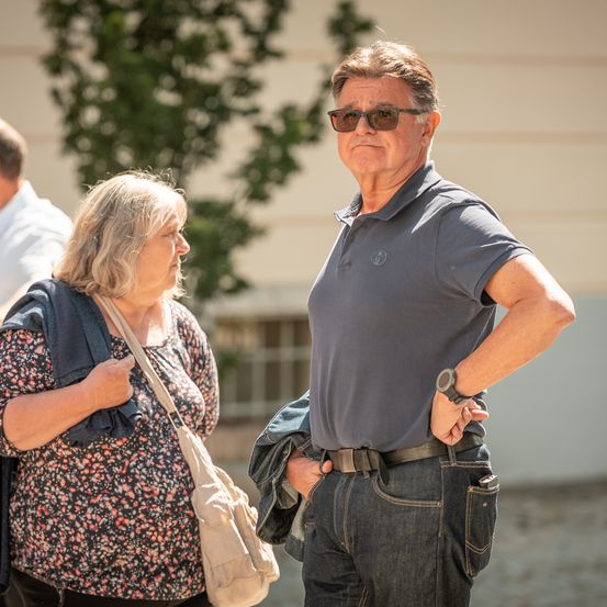 Ein älterer Mann und eine ältere Frau stehen draußen und sehen sich an. Der Mann trägt eine Sonnenbrille und ein graues Poloshirt, während die Frau ein Blumenoberteil trägt und eine Tasche bei sich hat. Dahinter ist ein Gebäude mit einem Fenster und einem Baum zu sehen.
