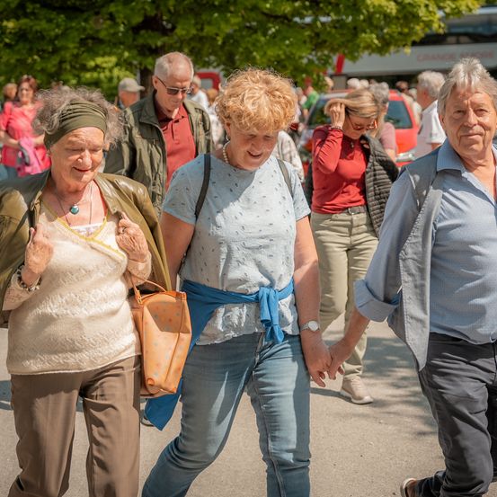 Eine Gruppe älterer Erwachsener geht draußen. Zwei Frauen und ein Mann halten sich an den Händen. Die Frau links trägt eine Stirnband und eine Halskette. Die Frau in der Mitte trägt einen blauen Schal. Der Mann trägt eine Uhr.