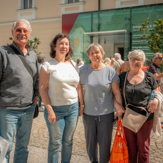 Vier Erwachsene, zwei Männer und zwei Frauen, stehen gemeinsam im Freien vor einem Gebäude mit einer Glaswand. Sie lächeln für ein Foto.