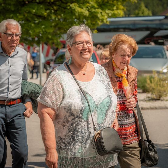 Drei ältere Menschen gehen auf einer Straße. Eine Frau mit einem Schmetterlings-gedruckten Oberteil lächelt. Ein älterer Mann mit Brille und Gürtel steht hinter ihr. Eine weitere ältere Frau mit Schal und Tasche schaut nach rechts.