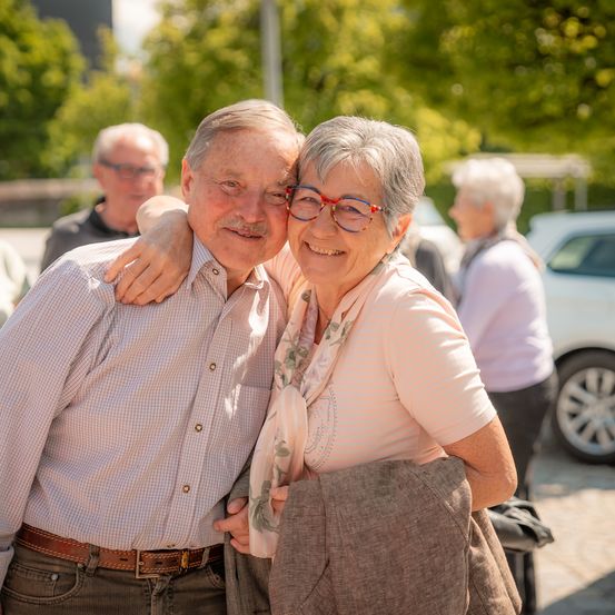 Ein älteres Paar steht lächelnd für ein Foto im Freien. Der Mann hat seinen Arm um die Frau gelegt, die eine Brille und einen Schal trägt. Sie befinden sich in einem sonnigen, grün bewachsenen Bereich mit einem geparkten weißen Auto im Hintergrund.