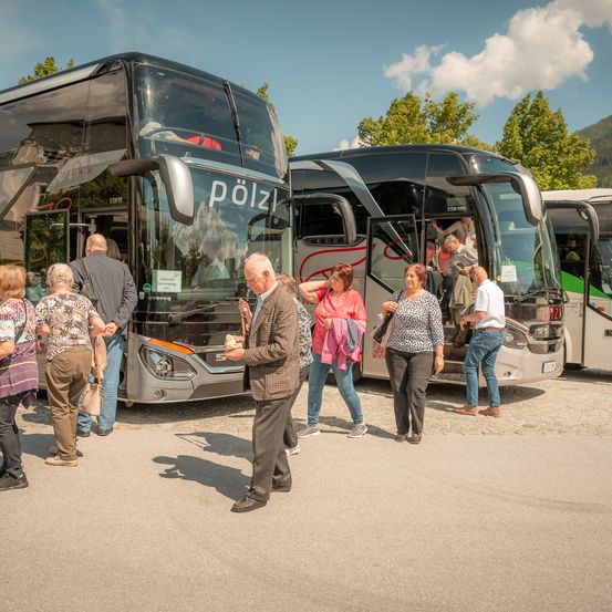 Eine Gruppe von Menschen steht draußen in der Nähe eines schwarzen Polzin-Tourbusses. Ein Mann spricht mit einem anderen Mann im Vordergrund. Einige Leute steigen in den Bus ein.