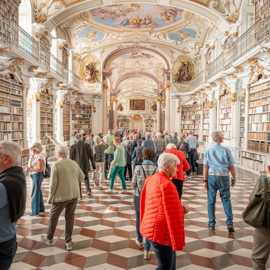 Eine Gruppe von Menschen geht in einer Bibliothek mit hohen Decken und kunstvollen Verzierungen. Die Bibliothek ist mit Bücherregalen gefüllt und die Menschen erkunden sie.