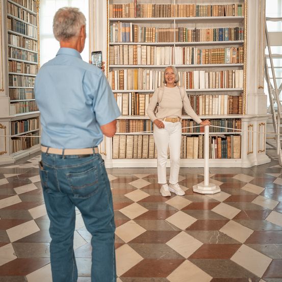 Ein älterer Mann fotografiert eine lächelnde ältere Frau hinter einer Seilbarriere in einer Bibliothek mit gefliesten Böden und Bücherregalen voller Bücher.