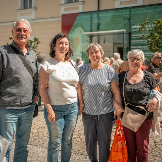 Vier Erwachsene stehen zusammen vor einem Gebäude mit einer Glaswand. Sie scheinen für ein Foto zu posieren.