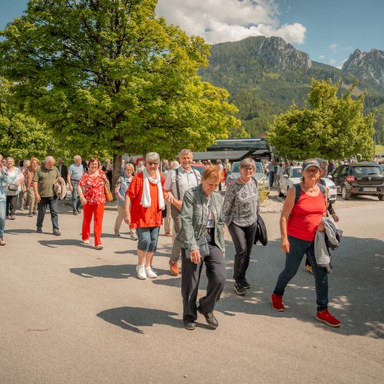 Eine Gruppe älterer Menschen geht auf einer Straße mit Bergen im Hintergrund. Einige tragen Taschen, und es sind geparkte Autos und ein Bus zu sehen.