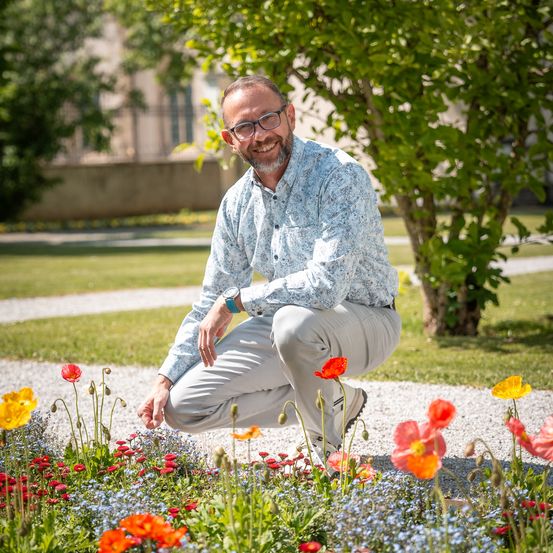 Ein Mann mit Brille kniet in einem Garten voller roter und gelber Blumen. Er trägt ein hellblaues Hemd und khakifarbene Hose. Der Hintergrund zeigt einen Rasenbereich mit Bäumen und einem Gebäude.