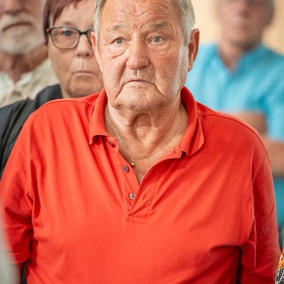 Ein älterer Mann in einem roten Polohemd steht mit ernster Miene, mit verschwommenen Gesichtern anderer im Hintergrund.