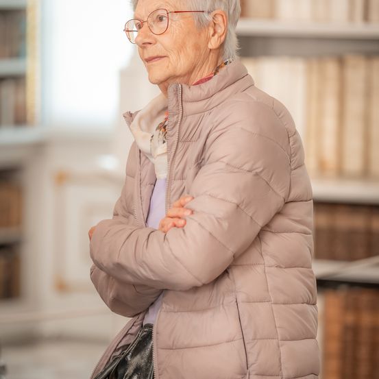 Eine ältere Frau mit Brille steht mit verschränkten Armen in einer Bibliothek. Sie trägt eine beige Daunenjacke und eine schwarze Handtasche.