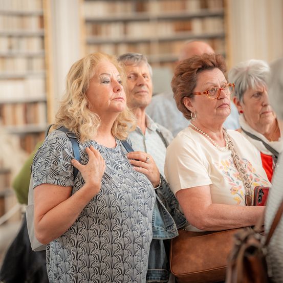 Mehrere ältere Personen stehen in einem Raum mit Bücherregalen und blicken nach vorne, vielleicht in einer Bibliothek.