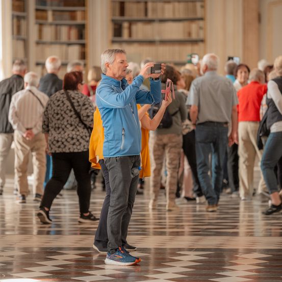 Ein älterer Mann in einem blauen Jackett macht ein Foto mit seinem Handy in einer Bibliothek. Mehrere Menschen gehen um ihn herum.