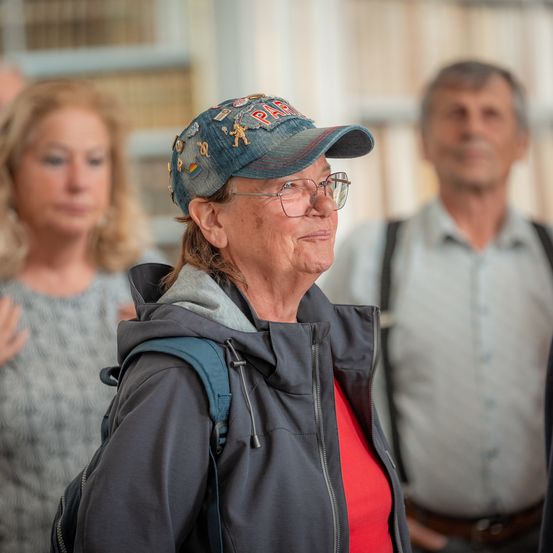 Eine ältere Frau mit einer Jeanskappe, Brille und einem roten Shirt steht in einem Raum mit zwei anderen Personen, von denen eine Hosenträger trägt.