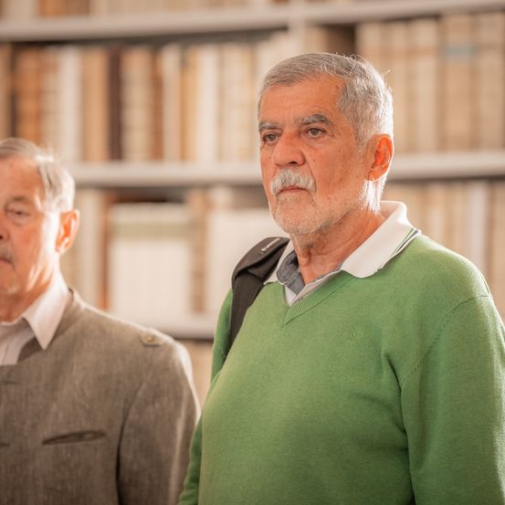 Zwei ältere Männer stehen vor einem Bücherregal voller Bücher. Einer trägt einen grünen Pullover mit einem Rucksack.