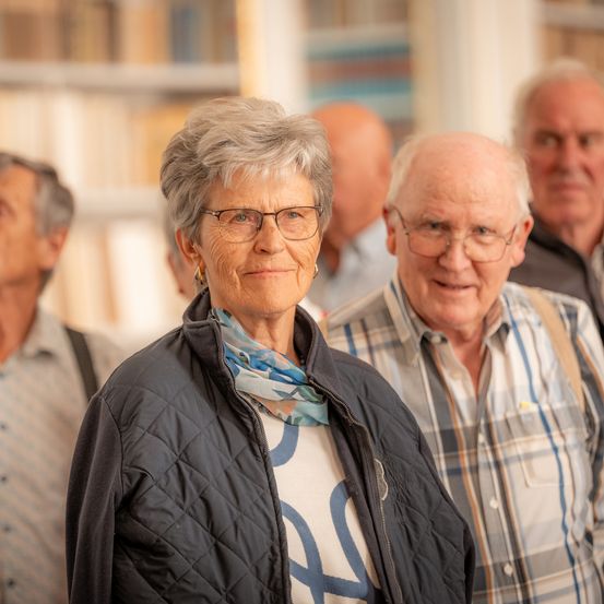 Eine ältere Frau mit Brille und Schal steht mit zwei älteren Männern in einem Raum mit Bücherregalen.