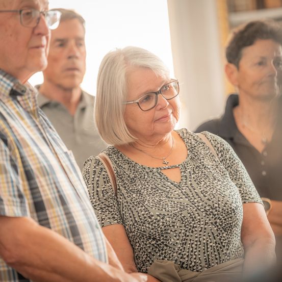 Eine ältere Frau mit Brille steht zwischen zwei Männern. Sie alle schauen nach links, vielleicht bei einer Veranstaltung oder Versammlung. Die Frau hat kurze weiße Haare und trägt eine Halskette.