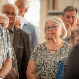 Eine Gruppe älterer Menschen steht in einem Raum. Eine ältere Frau mit Brille und Halskette schaut zur Seite.