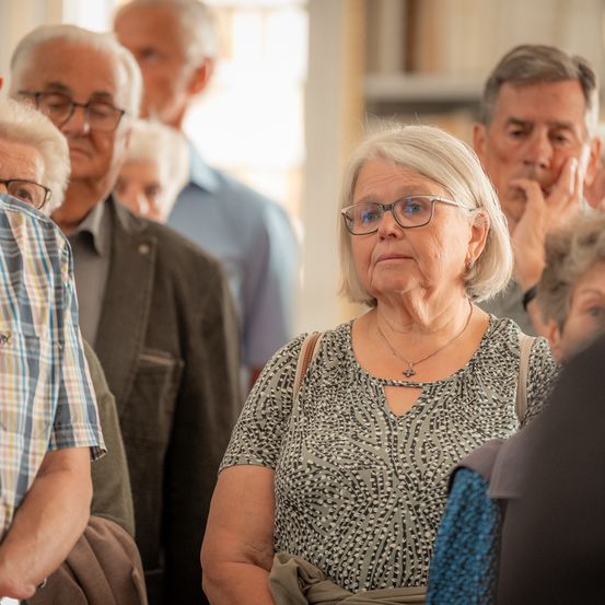 Eine Gruppe älterer Menschen steht in einem Raum. Eine ältere Frau mit Brille und Halskette schaut zur Seite.