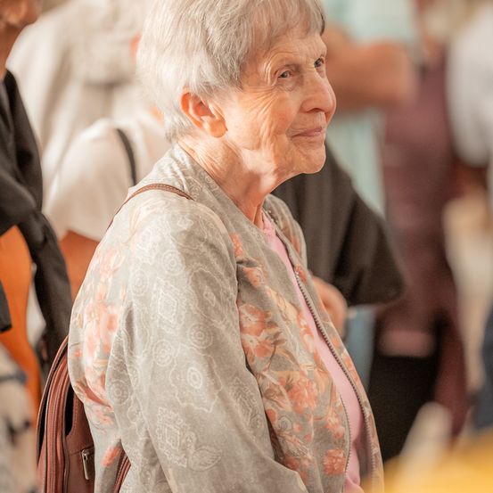Eine ältere Frau mit grauem Haar steht in einer Menschenmenge, lächelt und trägt eine Bluse mit Blumenmuster.