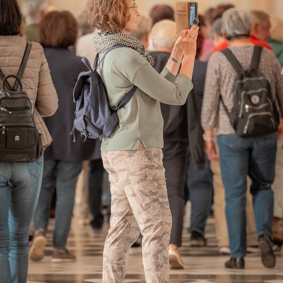 Eine ältere Frau geht in einer Menschenmenge und hält ihr Telefon in der rechten Hand, um ein Foto zu machen. Sie trägt einen Schal und einen Rucksack. Hinter ihr gehen Leute in verschiedene Richtungen.