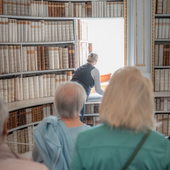 Eine Frau in einer Weste steht in einer Bibliothek mit hohen Regalen und schaut aus dem Fenster. Zwei ältere Frauen schauen sich die Bücher vor ihr an.