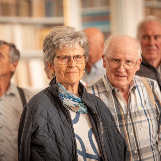Eine ältere Frau mit Brille und Schal steht zwischen zwei älteren Männern in einer Bibliothek.
