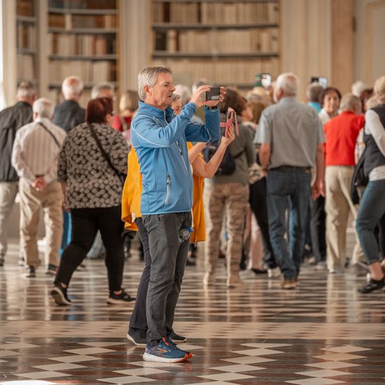 Ein älterer Mann macht ein Foto in einer Bibliothek, wo eine Menschenmenge herumläuft.