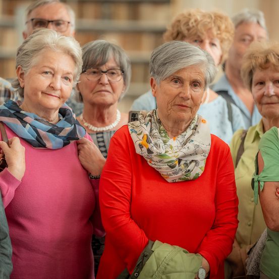 Eine Gruppe älterer Frauen steht zusammen. Die Frau vorne trägt ein rotes Shirt und einen Blumenschal. Sie scheinen an einer Veranstaltung teilzunehmen.