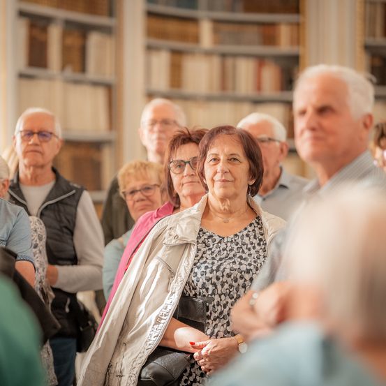 Eine Gruppe älterer Menschen steht in einer Bibliothek, trägt Brillen und hält ihre Sachen fest. Die Frau in der Mitte lächelt und schaut nach vorne.