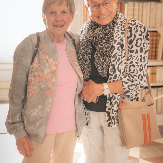Zwei ältere Frauen stehen lächelnd zusammen in einem Raum mit Bücherregalen im Hintergrund. Eine trägt eine Jacke mit Blumenmuster, die andere einen Leopardenschal.