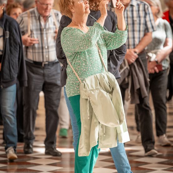 Eine ältere Frau hält einen Moment mit ihrer Kamera fest, während sie in einem gefliesten Flur unter einer Menschenmenge steht.