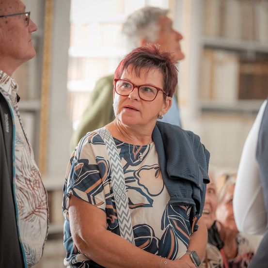 Eine ältere Frau mit kurzen roten Haaren und einer Brille steht und sieht nach vorne. Hinter ihr beobachtet ein Mann mit weißen Haaren und einem grünen Shirt. Im Hintergrund sind zwei weitere Personen teilweise zu sehen.