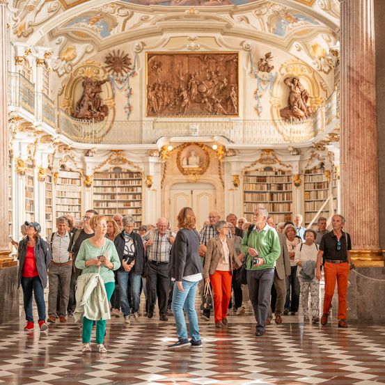 Eine Gruppe älterer Besucher erkundet eine reich verzierte Bibliothek mit hohen Decken und dekorativen Kunstwerken und geht durch einen gemusterten Boden.
