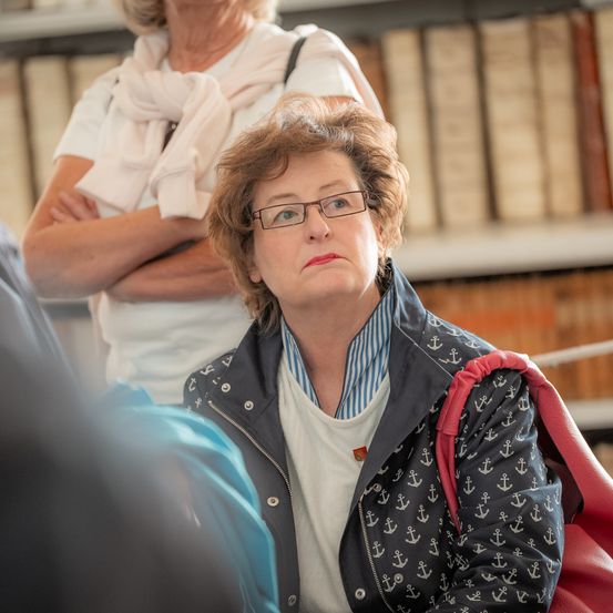 Eine Frau mit kurzen Haaren und Brille schaut in einer Bibliothek nach unten, sie trägt eine Jacke mit Ankermuster. Eine weitere Frau mit einem Schal steht hinter ihr, Arme verschränkt.