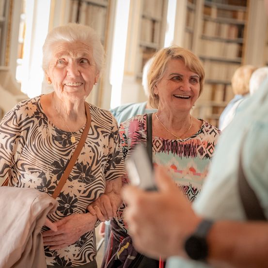 Zwei ältere Frauen lächeln und lachen, während sie zusammen in einer Bibliothek stehen, umgeben von Bücherregalen.