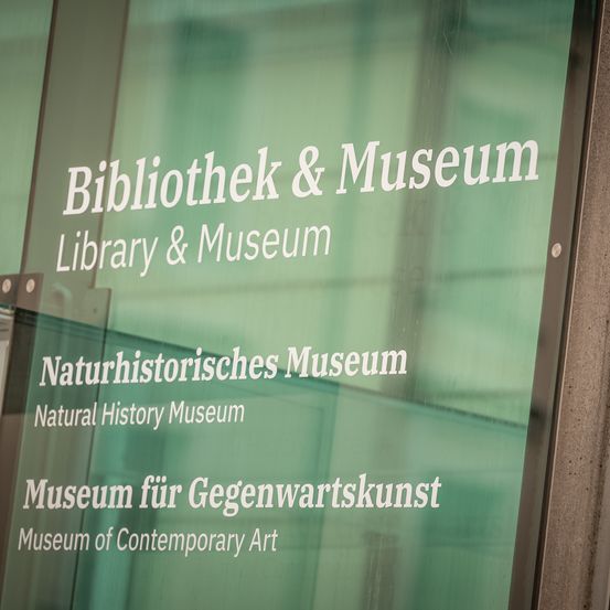 Ein Schild zeigt die Namen mehrerer Institutionen: Bibliothek & Museum, Naturhistorisches Museum und Museum für Gegenwartskunst.