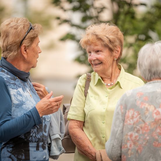Drei ältere Frauen unterhalten sich in einem Park. Die Frau auf der linken Seite trägt ein blaues Langarmshirt mit Blumenmustern, während die Frau in der Mitte ein gelbes Oberteil mit einer Halskette trägt. Die Frau auf der rechten Seite trägt ein Top mit Blumenmuster. Bäume und ein Gebäude sind im Hintergrund zu sehen.