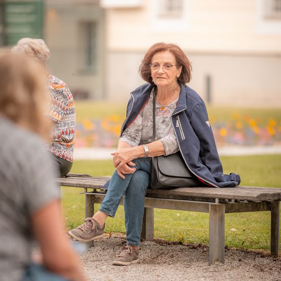 Eine ältere Frau mit Brille und blauer Jacke sitzt auf einer Parkbank und schaut nachdenklich. Hinter ihr beobachtet sie eine andere Frau in einem gemusterten Shirt.