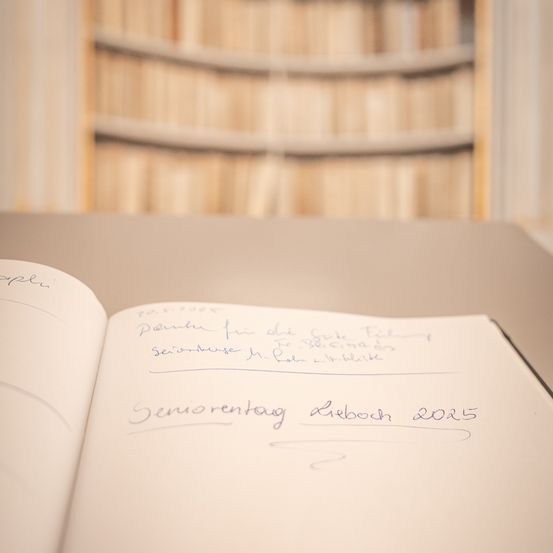 Ein offenes Notizbuch mit handgeschriebenem Text mit dem Titel 'Seniorentag Lieboch 2025' und anderen Notizen liegt auf einem Tisch mit einem verschwommenen Bücherregal im Hintergrund.