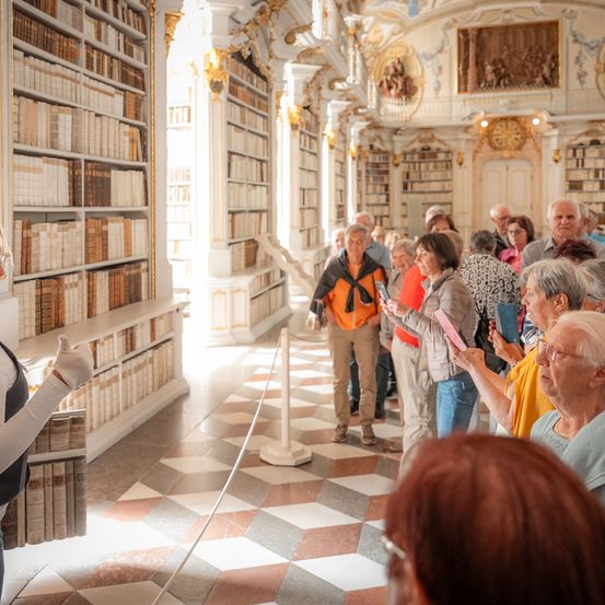 Eine Gruppe von Menschen ist in einer Bibliothek. Eine Frau hält ein Buch und eine andere zeigt. Sie sind von Bücherregalen umgeben.