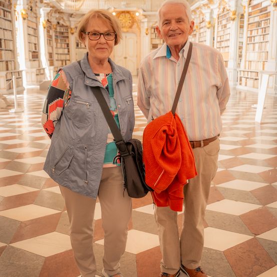 Ein älteres Paar steht in einer Bibliothek mit gemusterten Böden. Die Frau trägt eine Brille und eine Weste, während der Mann ein gestreiftes Hemd und einen roten Schal über der Schulter hat.