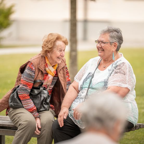 Zwei ältere Frauen sitzen auf einer Parkbank und unterhalten sich lächelnd. Eine Frau trägt eine Brille und eine Halskette. Die andere hat eine Handtasche bei sich. Hinter ihnen ist ein Baum und ein verschwommener Hintergrund zu sehen.