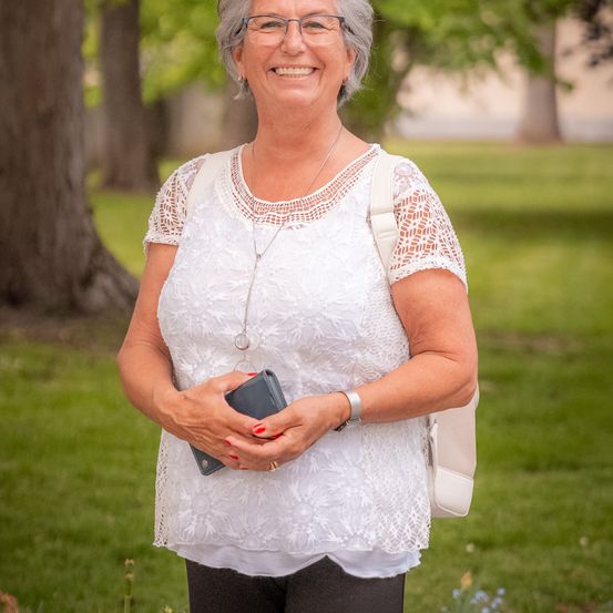 Eine lächelnde ältere Frau in einem weißen Blusenoberteil mit Spitzenbesatz steht in einem Park, hält eine Geldbörse und trägt eine Brille, eine Halskette und eine Uhr.