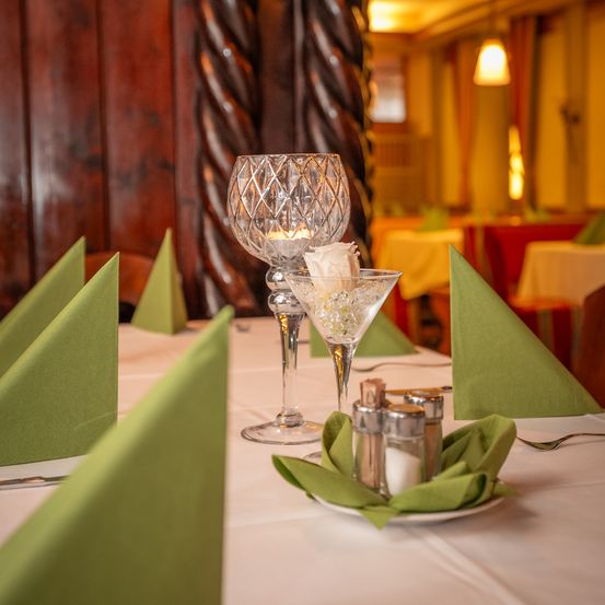 Ein Restauranttisch ist mit grünen Servietten, Weingläsern und Gewürzen gedeckt. Im Hintergrund befinden sich Holzwände und eine Spiralsäule.