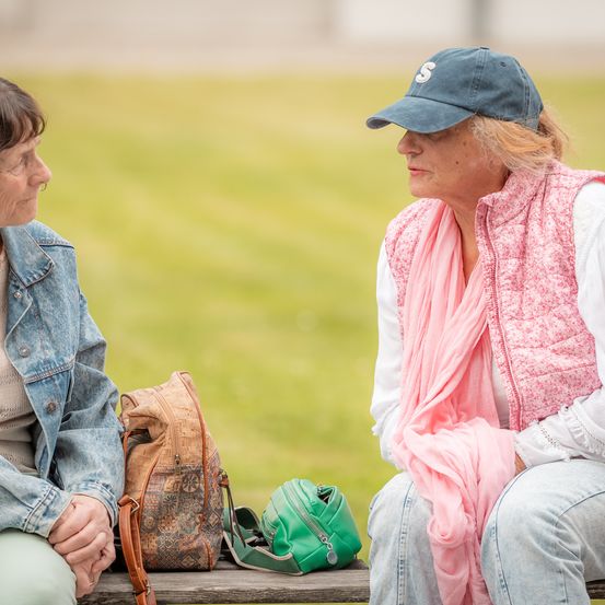 Zwei Frauen sitzen auf einer Bank mit Taschen in der Nähe, eine Frau trägt eine Jeansjacke und die andere eine rosa Weste und eine blaue Kappe.