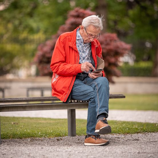 Ein älterer Mann sitzt auf einer Parkbank im Park, trägt eine Brille und eine rote Jacke. Er hält einen Geldbeutel und schaut auf sein Handy.