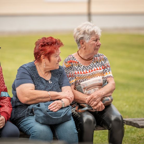 Zwei ältere Frauen sitzen auf einer Parkbank und unterhalten sich. Eine trägt ein blaues Oberteil und Jeans, die andere ein orangefarbenes gemustertes Oberteil und Lederhosen. Beide halten Taschen.