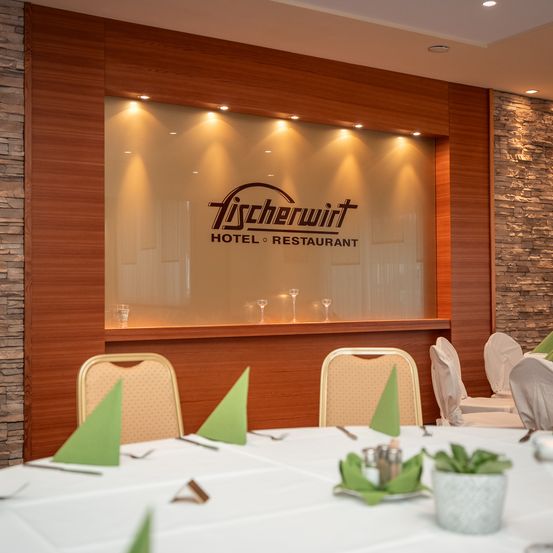 Ein Restaurant-Essbereich mit einer großen Glaswand, die den Namen Tischlerwirt zeigt. Holzwände und Steinakzente schmücken den Raum. Tische sind mit weißen Tüchern, Besteck und grünen Servietten gedeckt. Weingläser werden auf der Glaswand reflektiert.