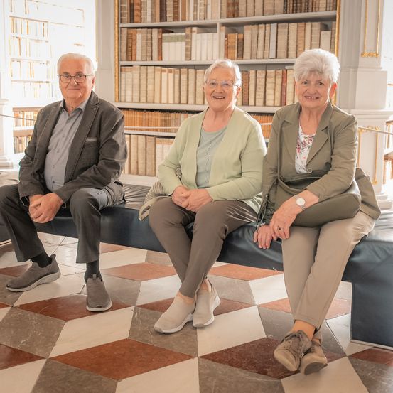 Drei ältere Personen sitzen auf einer Bank, lächeln und schauen in die Kamera. Dahinter befinden sich hohe Bücherregale voller Bücher.