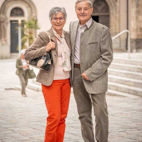 Ein älteres Paar steht lächelnd auf einer Kopfsteinpflasterstraße. Die Frau trägt orangefarbene Hosen und eine leichte Jacke, während der Mann einen grauen Anzug trägt. Hinter ihnen führen Stufen zu einer Rundbogentür.