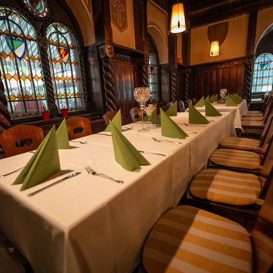 Ein langer Esstisch mit Besteck und Servietten. Der Tisch ist mit einer weißen Tischdecke bedeckt. Die Stühle sind mit gestreiftem Stoff bezogen. Der Raum hat Holzwände und Buntglasfenster.